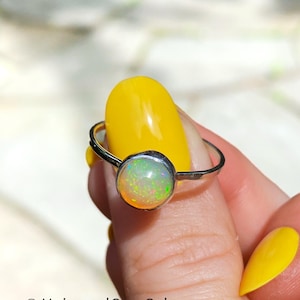 May include: A silver ring with a round opal gemstone. The opal displays a mix of colors, including green, yellow, and orange. The ring is held up against a yellow background.