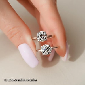 May include: Two silver engagement rings with round brilliant cut diamonds. The rings are stacked on top of each other, with the larger diamond on top. The diamonds are clear and sparkly.