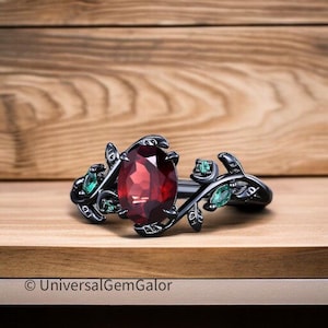 以下が含まれることがあります： 大きな楕円形の赤い宝石と小さな緑色の宝石をあしらった黒い指輪。葉とつるのフローラルデザイン。木製の表面に指輪が飾られています。