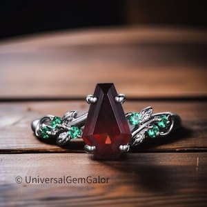 May include: A silver ring with a coffin-shaped, dark red gemstone as the focal point. The ring is embellished with green gemstones and leaf motifs. The text "UniversalGemGalor" is visible.