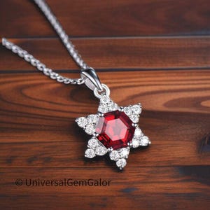 May include: A silver necklace with a star-shaped pendant. The pendant features a large, faceted, red gemstone at the center, surrounded by smaller, clear gemstones. The necklace is displayed on a wooden surface.