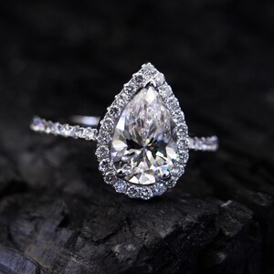 May include: A pear-shaped diamond engagement ring with a halo setting. The ring is set in white gold and features a large, sparkling diamond in the center.
