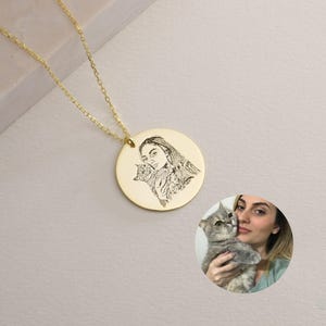 May include: A gold-coloured necklace with a circular pendant. The pendant features a detailed engraved portrait of a person holding a cat. The chain is delicate, and the pendant is the focal point of the jewellery.