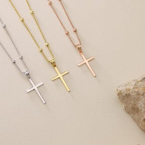 May include: Three gold cross necklaces with different chain styles. The necklaces are on a light beige background.