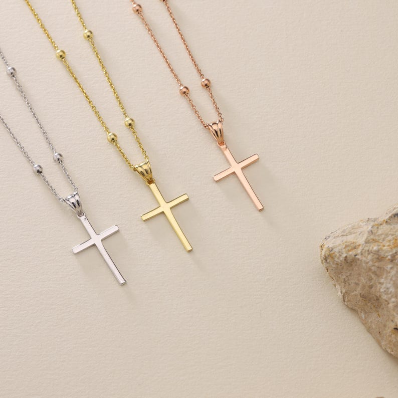 May include: Three gold cross necklaces with different chain styles. The necklaces are on a light beige background.