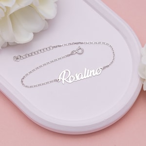 May include: A silver bracelet inscribed with the name "Rosaline" in a cursive script. The bracelet rests on a white tray, set against a pink backdrop, with white flowers.