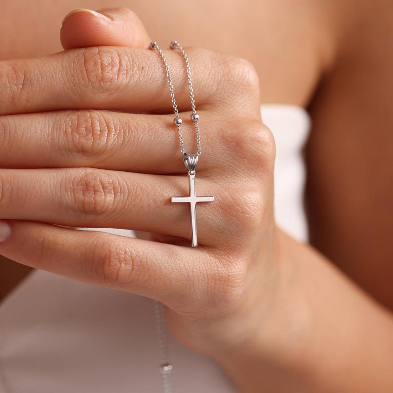 May include: A silver chain necklace with a small silver cross pendant. The necklace is held in a person's hand.