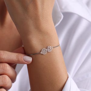 May include: A silver bracelet featuring two delicate floral charms. One charm is a detailed openwork design, and the other is a rose. The bracelet is on a fine chain, and is worn on a wrist.