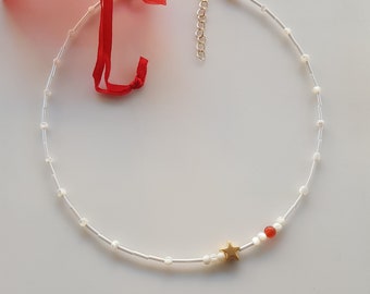 Puede incluir: Un delicado collar de cuentas blancas con un dije de estrella dorada y una sola cuenta roja. El collar está sobre un fondo blanco.