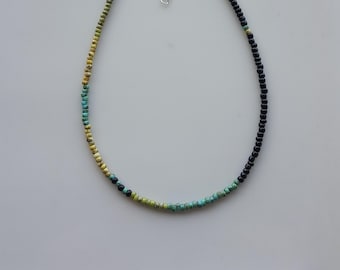 May include: A beaded necklace with a gradient design. The necklace features small, round beads in black, yellow, turquoise, and green. The necklace has a silver-colored clasp and is displayed on a white surface.