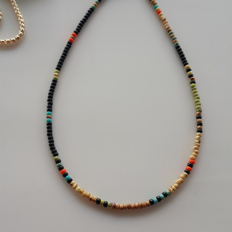 May include: A delicate beaded necklace with black, beige, green, and orange beads. The necklace is made of small, round beads and has a simple, minimalist design.