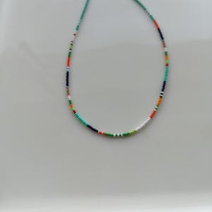 Puede incluir: Un collar de cuentas de colores con un cordón turquesa. El collar presenta pequeñas cuentas redondas en una variedad de colores, incluyendo rojo, naranja, verde, blanco y azul marino. Las cuentas están ensartadas para crear un accesorio delicado y llamativo.