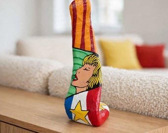May include: A vertical, abstract sculpture featuring a painted profile of a person with blonde hair. The top section has orange and red vertical stripes, the middle shows a green background, and the base displays a blue section with a yellow star.