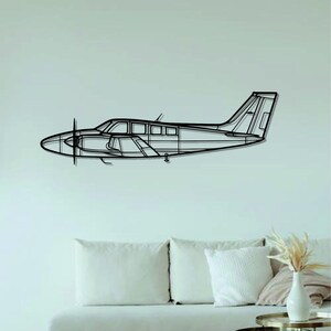 May include: A black metal outline of a small airplane, hanging on a white wall above a white couch with pillows. The couch is in front of a wooden floor with a woven rug and two gold metal side tables.