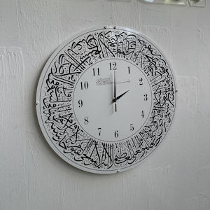 May include: A white round wall clock with black Arabic calligraphy around the edge and black numerals. The clock hands are black. The word "DESIGN" is in neon lights above the clock.