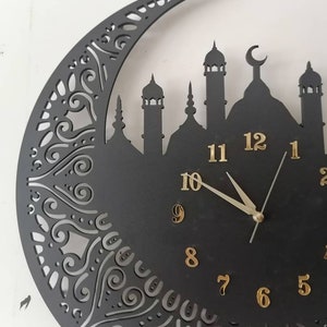 May include: Black clock with gold numbers and hands, shaped like a crescent moon with a mosque silhouette. The clock has a decorative design around the edge.