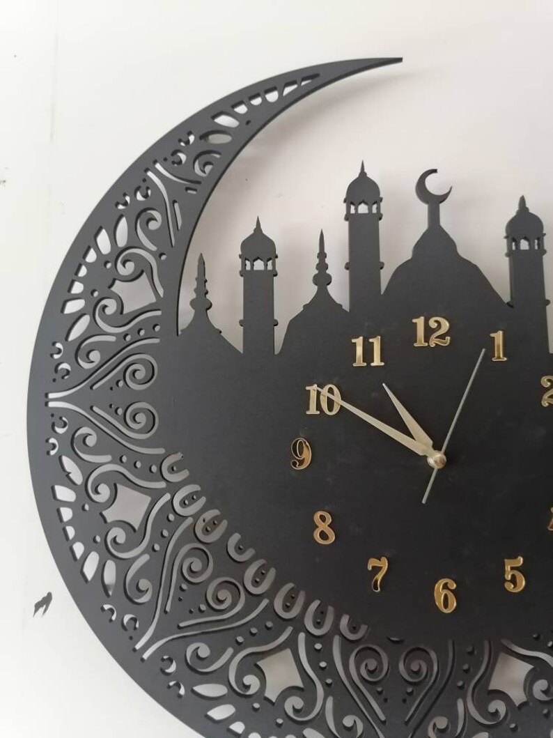 May include: Black clock with gold numbers and hands, shaped like a crescent moon with a mosque silhouette. The clock has a decorative design around the edge.