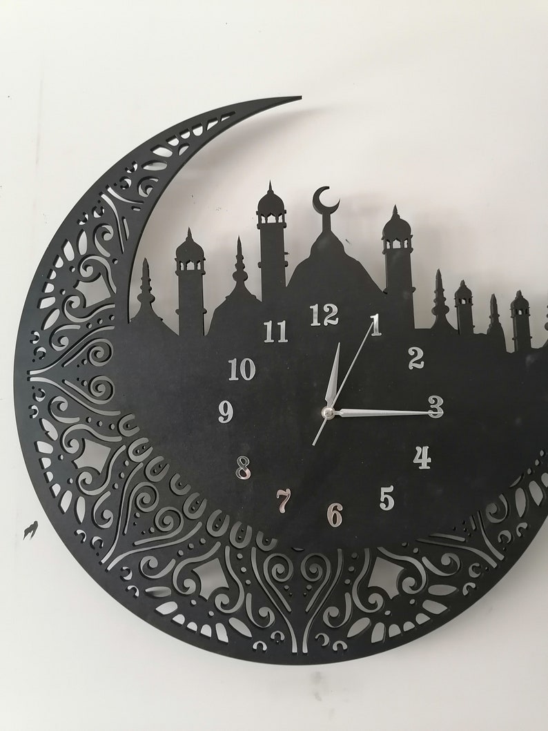 May include: Black wall clock with a crescent moon and mosque silhouette design. The clock face has Roman numeral markings and silver hands.