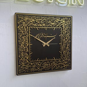 Peut inclure: Horloge murale carrée noire avec calligraphie arabe dorée sur le pourtour et sur le cadran. Les aiguilles de l'horloge sont dorées. L'horloge est fixée sur un mur blanc. Le mot "DESIGN" est en néon au-dessus de l'horloge.