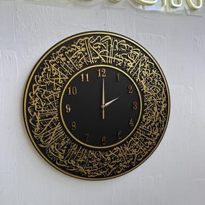 Può includere: Un orologio da parete rotondo nero con calligrafia araba dorata sul bordo e numeri dorati. Le lancette dell'orologio sono dorate. L'orologio è montato su una parete bianca.