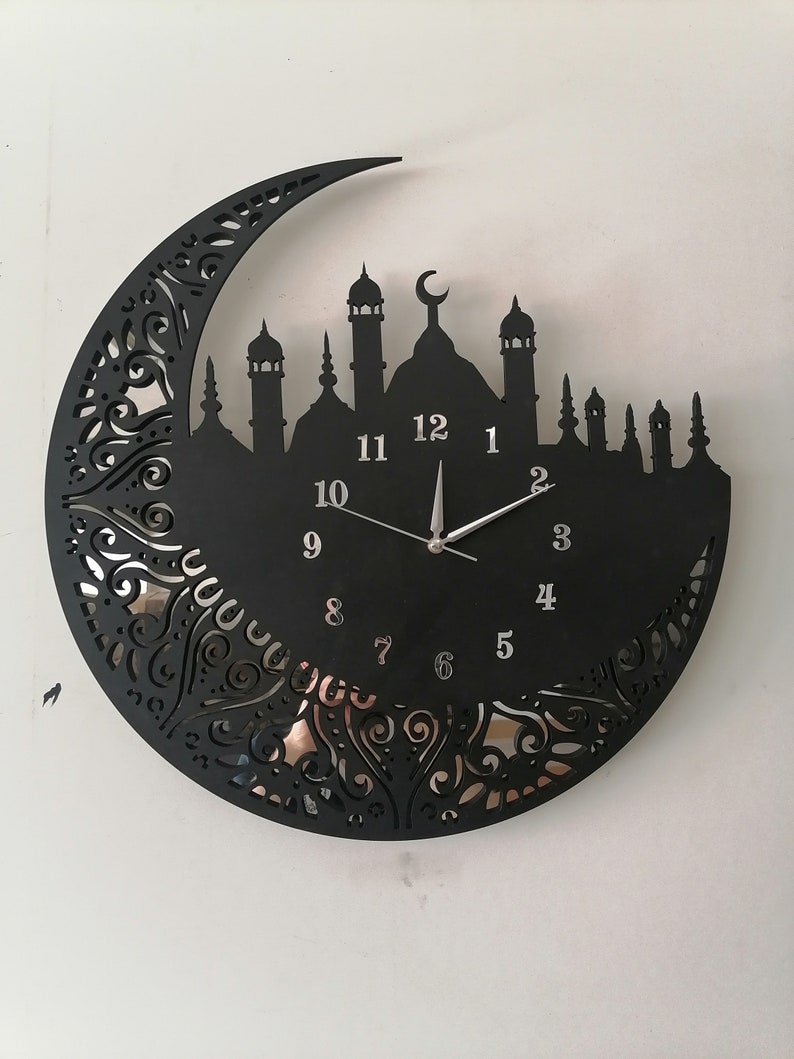 May include: Black metal wall clock with a crescent moon design and a silhouette of a mosque. The clock face features Arabic numerals and a silver hand.