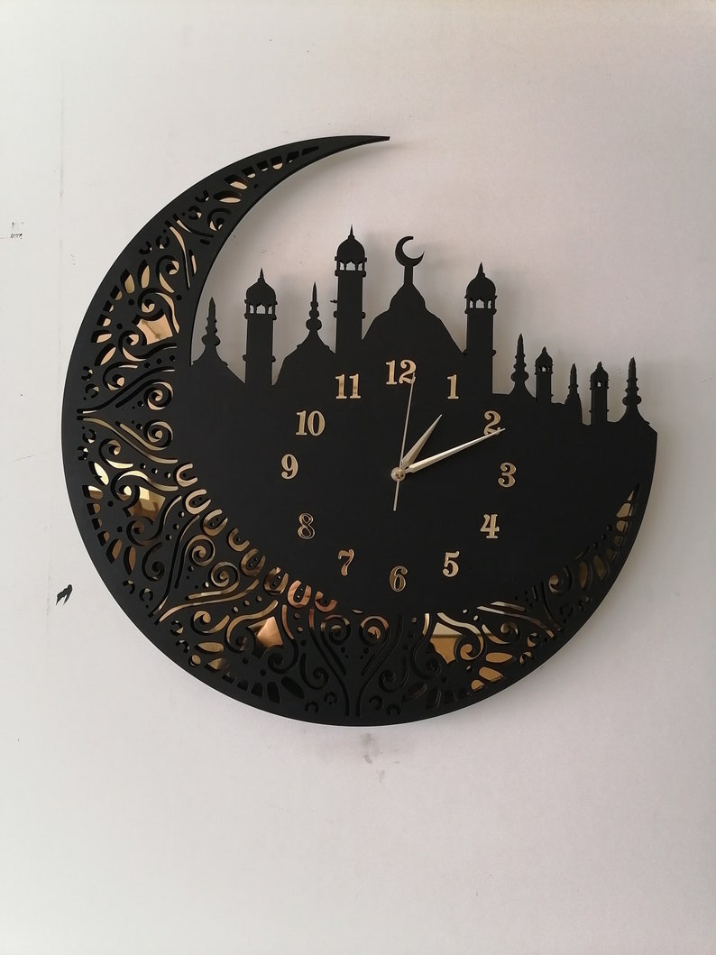 May include: Black wall clock with a crescent moon and mosque silhouette design. The clock face has gold Arabic numerals and a gold hour and minute hand.