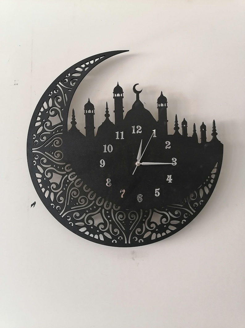 May include: Black metal wall clock with a crescent moon and mosque silhouette design. The clock face has Roman numeral markings and a white hour and minute hand.
