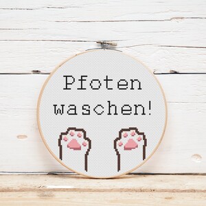 Op de afbeelding: Een witte borduurring met de tekst "Pfoten waschen!" en twee bruine poot afdrukken met roze voetkussens.