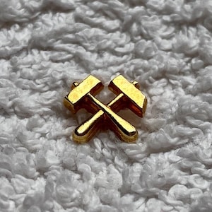 May include: A small, gold-coloured pin featuring a crossed hammer design. The pin has a shiny, reflective surface and is set against a textured, white background. The design is simple and likely represents a trade or profession.
