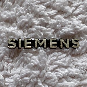 Può includere: Una piccola spilla color argento con la scritta "SIEMENS" in maiuscolo. La spilla ha un bordo nero ed è posta su uno sfondo bianco strutturato.