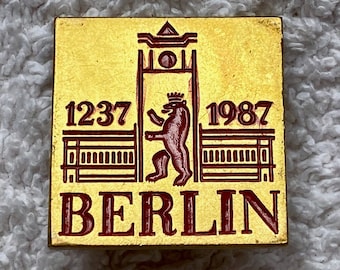 Puede incluir: Un pin de metal dorado con un diseño rojo y blanco. El pin presenta una imagen estilizada de una puerta con un oso de pie en el medio. El texto "1237" y "1987" están a cada lado de la puerta. El texto "BERLIN" está en la parte inferior del pin.