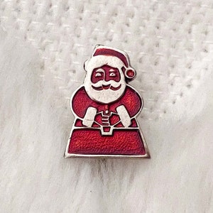 Pode incluir: Um broche de esmalte vermelho e branco com um Papai Noel de desenho animado segurando um saco de presentes.