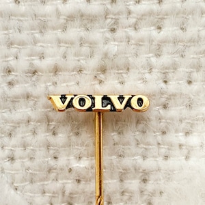 Può includere: Spilla dorata con la scritta "VOLVO" in lettere nere. La spilla ha un lungo e sottile stelo ritorto. Lo sfondo è un tessuto strutturato bianco sporco.