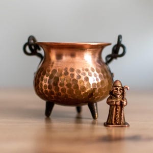 May include: A small copper cauldron with a hammered texture, featuring a handle and three legs. Beside it stands a miniature figurine of a wizard or gnome. The items are displayed on a wooden surface.