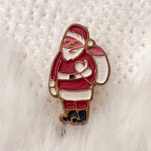 Pode incluir: Um alfinete de esmalte do Papai Noel com uma borda dourada. O alfinete apresenta um terno vermelho, barba branca e um saco rosa. O alfinete está sobre um fundo branco texturizado.