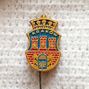 Puede incluir: Un pin de color dorado con un diseño de esmalte rojo, blanco y azul. El pin presenta un escudo con tres torres y la palabra "KRAKOW" sobre el escudo.