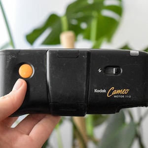 May include: A black Kodak Cameo Motor 110 film camera with an orange button. The camera has a flash and a small window on the top.