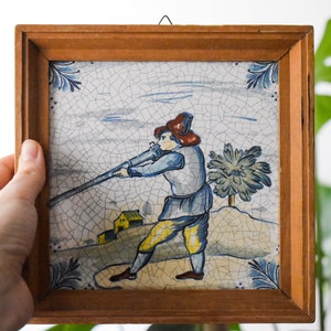 Könnte beinhalten: Eine gerahmte Fliese, die einen Mann in einem roten Hut zeigt, der eine lange Waffe hält, mit einem blauen und weißen Hintergrund und einem kleinen gelben Haus in der Ferne.