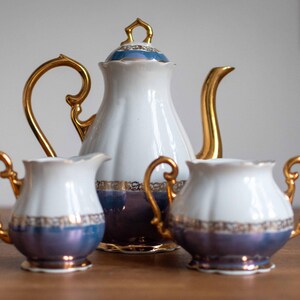May include: A white porcelain tea set with gold accents and a blue and purple ombre design. The set includes a teapot, creamer, and sugar bowl.