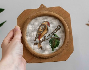 May include: A small, round, framed cross-stitch featuring a yellow bird perched on a branch with green leaves. The frame is made of light brown wood and has a slightly octagonal shape.