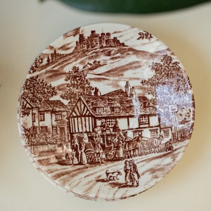 Könnte beinhalten: Eine braune und weiße Keramik-Untertasse mit einem Vintage-Design, das eine Stadtszene mit einem Schloss im Hintergrund zeigt. Die Untertasse hat einen braunen Rand und eine weiße Mitte mit einem braunen Design.