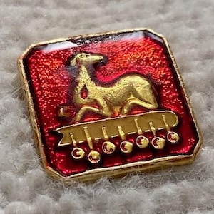 Op de afbeelding: Een kleine, rode emaille-pin met een gouden rand. De pin heeft een gestileerd beeld van een hert dat op een platform staat met een gouden lint en vijf gouden cirkels.