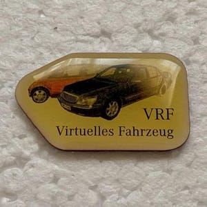 Può includere: Una piccola spilla rettangolare con sfondo giallo. Presenta due illustrazioni di auto, una arancione e una nera, con il testo "VRF Virtuelles Fahrzeug" sottostante. La spilla è probabilmente un oggetto da collezione o promozionale.
