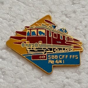 May include: A colorful enamel pin featuring a red and brown train with the text "SBB CFF FFS Re 4/4 I". The pin has a gold border and a blue and yellow background.