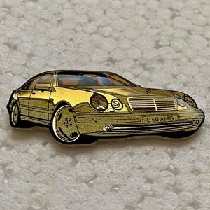 Puede incluir: Un pin dorado que representa un coche E 50 AMG clásico. El pin presenta ilustraciones detalladas de la parte delantera, lateral y ruedas del coche. El texto "E 50 AMG" es visible en la parte delantera del coche.