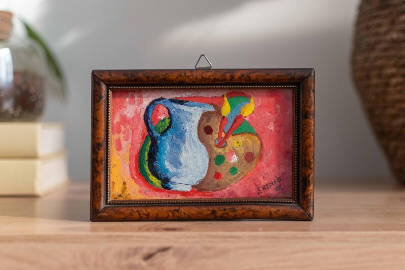May include: A framed painting of a blue pitcher and a paint palette with colorful paint splatters on a red background. The painting is signed "S. Kenis 1977".