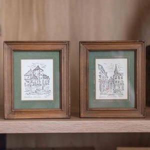 May include: Two small rectangular wooden frames with dark brown finish, each containing a black and white ink sketch of a European town scene. The sketches are set against a textured green matting with a thin gold border inside the frame.