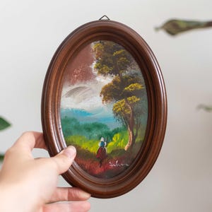 May include: A small oval framed painting of a landscape scene with a single figure in the foreground. The painting is set against a backdrop of green hills, a blue sky, and a large tree. The frame is dark brown wood.