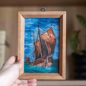 以下が含まれることがあります： 青い空と青い海に浮かぶ茶色の帆を張った帆船が描かれた、明るい色の木製フレームに入った小さな長方形の絵画。作品の上部には小さな金属製の吊り下げ用ループがあり、1982年という年号と署名が記されています。