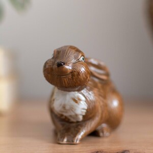 Może przedstawiać: Ceramiczna figurka brązowego królika z białym brzuchem i lekko złośliwym wyrazem twarzy.